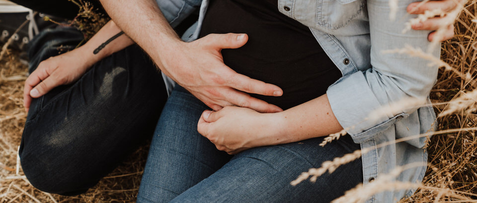 Schwangeres Pärchen sitzt in einem Feld und halten Hände an Babybauch Hochzeitsfotografin Eva Mertzen