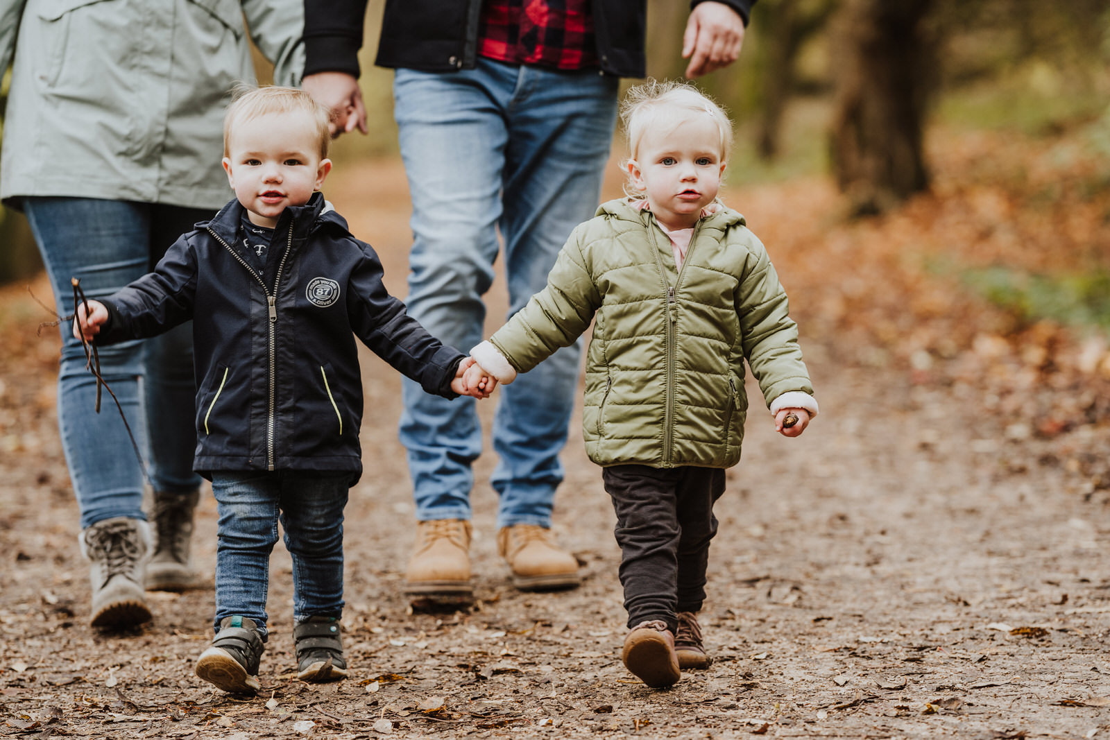 Zwei kleine Kinder gehen einen Waldweg entlang vor den Eltern und halten Händchen.