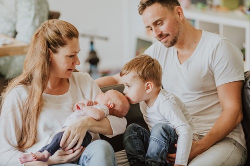 Ein kleiner Junge sitzt bei einem Mann auf dem Schoß und küsst ein Baby auf dem Kopf, dass von einer Frau gehalten wird.