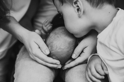 Ein kleiner Junge küsst ein Baby auf dem Kopf während das Baby bei den Eltern auf dem Schoß liegt.
