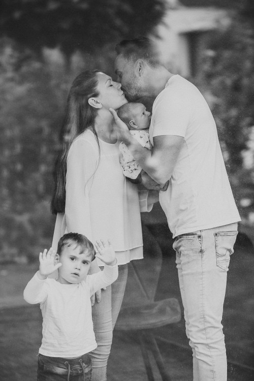 Ein Mann küsst eine Frau während er ein kleines Baby auf dem Arm hält und ein kleiner Junge vor der Frau steht.