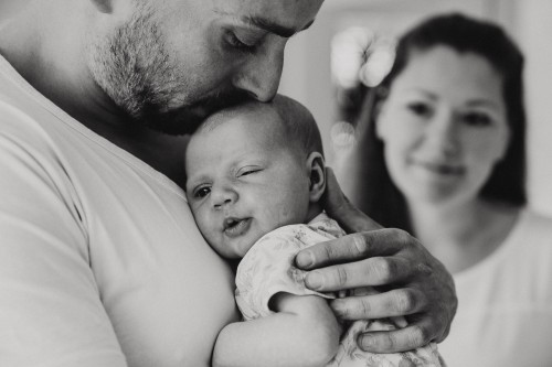 Ein Mann hält ein Baby und gibt ihm einen Kuss auf dem Kopf während eine Frau im Hintergrund zuschaut.