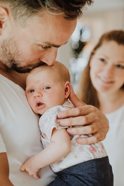 Ein Mann hält ein Baby und gibt ihm einen Kuss auf dem Kopf während eine Frau im Hintergrund zuschaut.