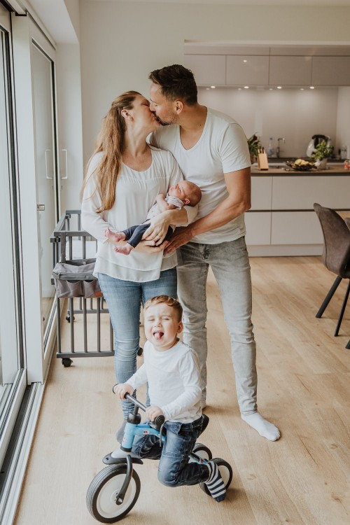 Eine Frau und ein Mann küssen sich während die Frau ein Baby auf dem Arm hält und ein kleiner Junge auf einem Dreirad sitzt.