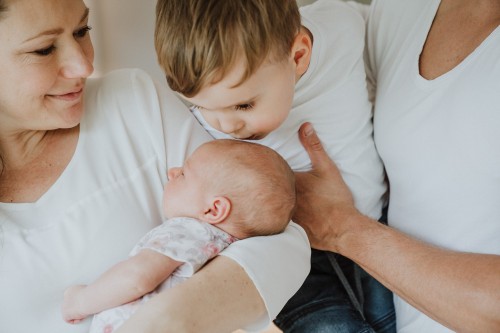Ein kleiner Junge wird von einem Mann gehalten und küsst ein Baby auf dem Kopf, dass eine Frau auf dem Arm hält.