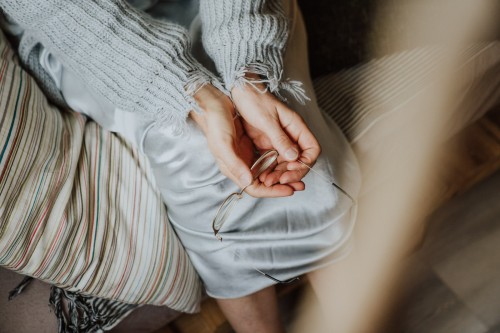 Eine Frau sitzt auf einem Bett und hält ihre Brille in den Händen.