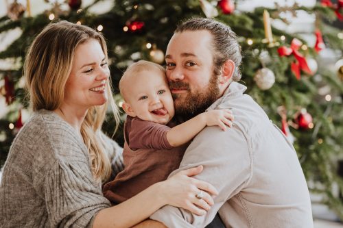 Eine Frau und ein Mann sitzen mit einem Baby vor dem Weihnachtsbaum.