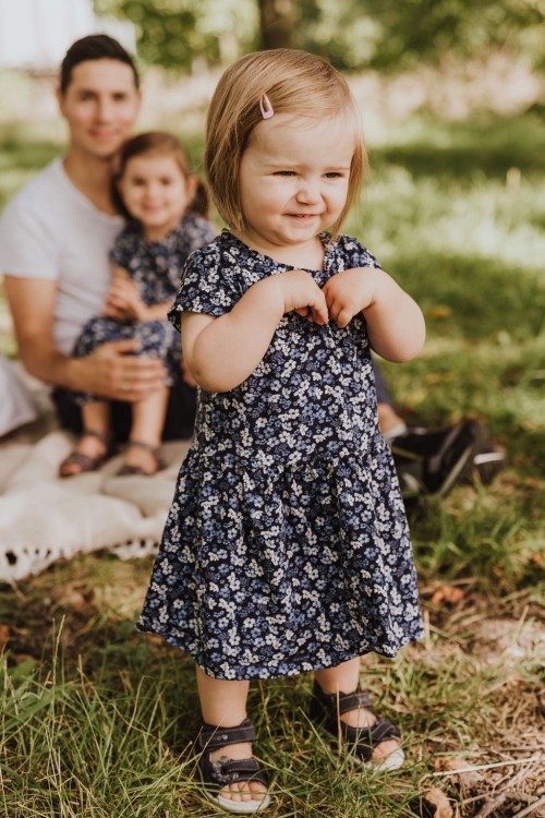Ein kleines Mädchen steht auf einer Wiese, dahinter sitzt ein Mann und hält ein kleines Mädchen auf dem Schoß.
