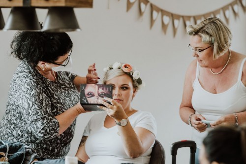 Eine Frau schminkt eine Braut und eine andere Frau schaut zu.