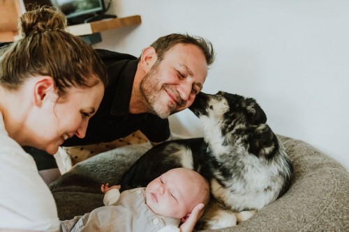 Ein Mann wird von einem Hund geleckt und eine Frau hält ein kleines Baby auf dem Arm.