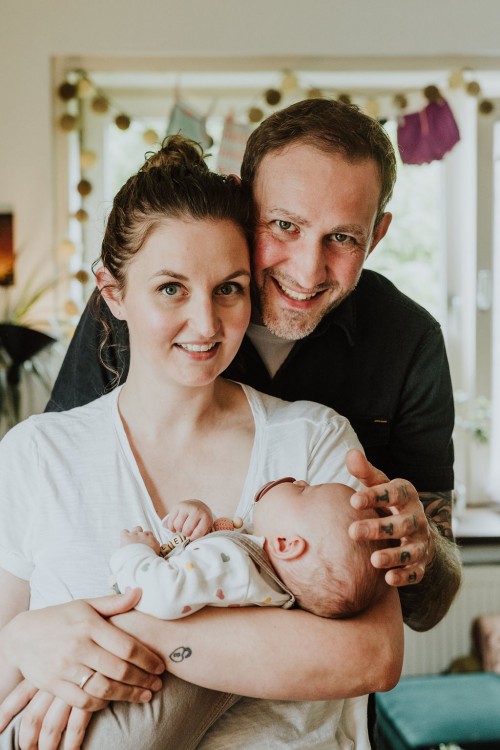 Eine Frau hält ein Baby auf dem Arm und ein Mann steht hinter den beiden und umarmt die Frau.