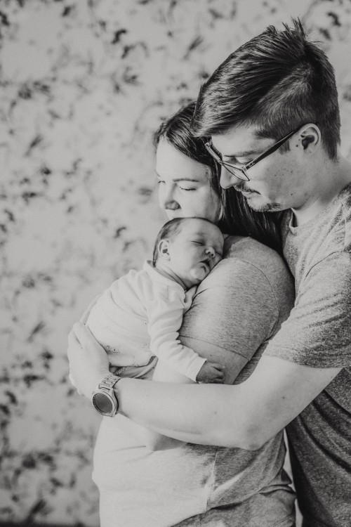 Ein kleines Baby schläft bei einer Frau auf dem Arm und ein Mann steht hinter der Frau und umarmt sie.