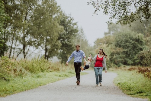 Ein Mann und eine Frau gehen einen Waldweg entlang und heben ein kleines Kind zusammen hoch.