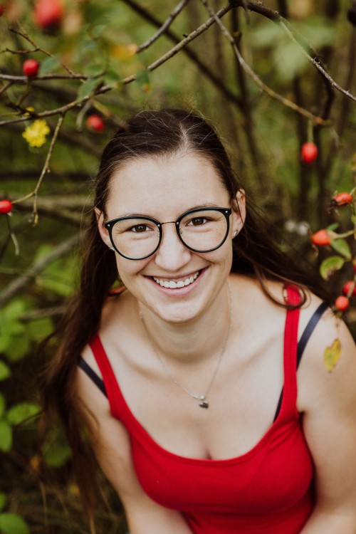 Eine Frau mit Brille sitzt im Wald und lächelt in die Kamera.