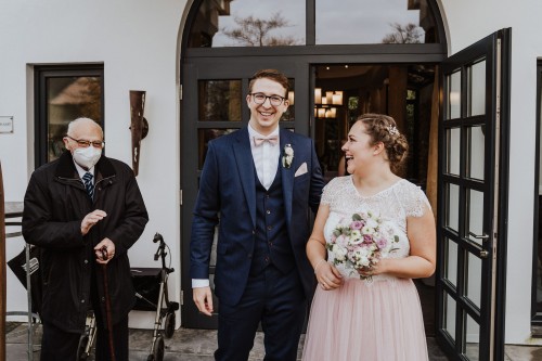 Ein Brautpaar steht vor einem Gebäude neben einem älteren Mann mit Atemschutzmaske.