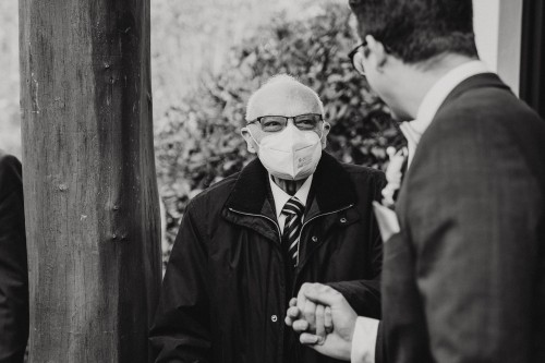 Ein älterer Mann mit einer Atemschutzmaske hält den Bräutigam an der Hand.