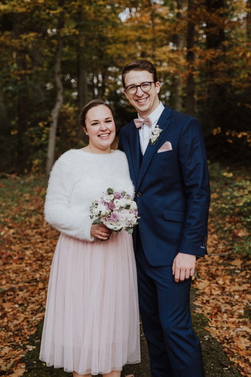 Eine Braut und ein Bräutigam stehen im Wald und schauen in die Kamera.