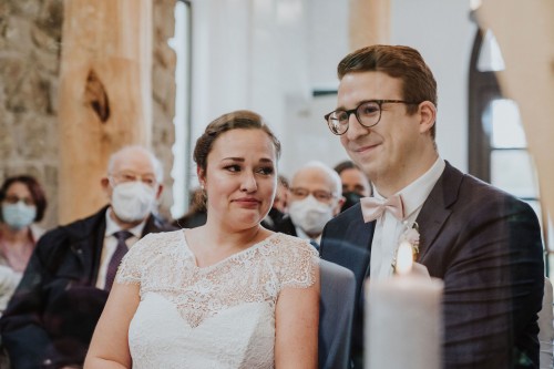 Eine Braut schaut zu dem Bräutigam hin, im Hintergrund sitzen Gäste.