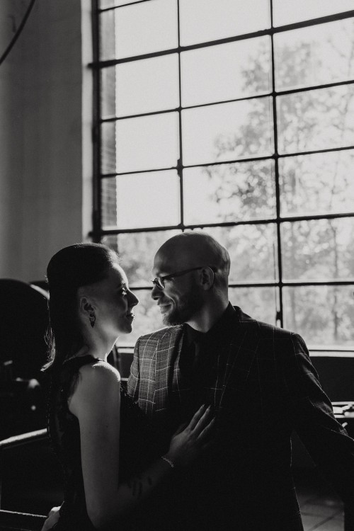 Ein schwarz-weißes Bild vom Brautpaar das sich vor einem großen Fenster umarmt.