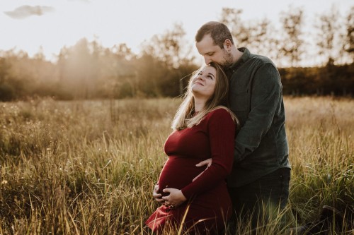 Ein Mann und eine Frau umarmen sich auf einem Feld und halten ihre Hände auf dem Babybauch.