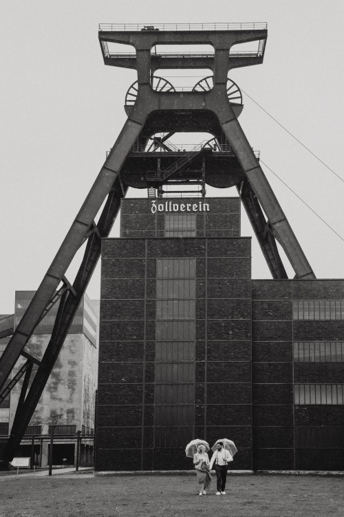 Ein Brautpaar steht mit Regenschirmen vor der Zeche Zollverein.