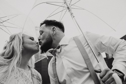 Ein schwarz-weißes Bild von einem Brautpaar dass sich zwischen den Regenschirmen küsst.