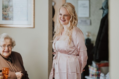 Eine Frau lacht herzlich in die Kamera während vor ihr eine ältere Dame sitzt.