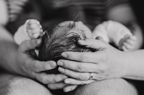 Ein kleines Baby liegt auf dem Schoß des Vaters und wird von Händen am Kopf festgehalten.