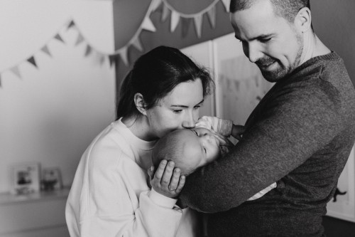 Ein Mann hält ein Baby im Arm und eine Frau küsst das Baby am Kopf.