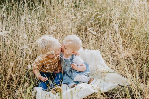 Zwei kleinen Kinder sitzen auf einer Decke auf einer Wiese und geben sich ein Küsschen.