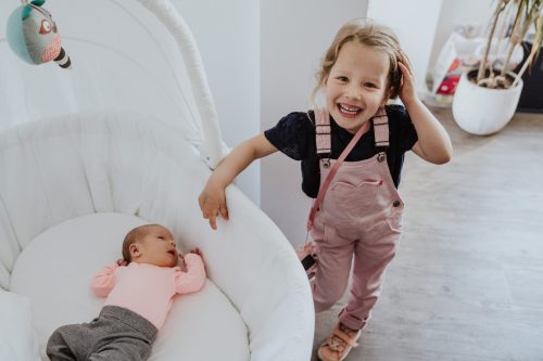 Ein kleines Mädchen steht neben einem Babybett mit kleinem Baby.