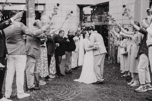 Ein Brautpaar küsst sich vor einem Eingang und die Gäste stehen Spalier und halten Rosen hoch in die Luft.