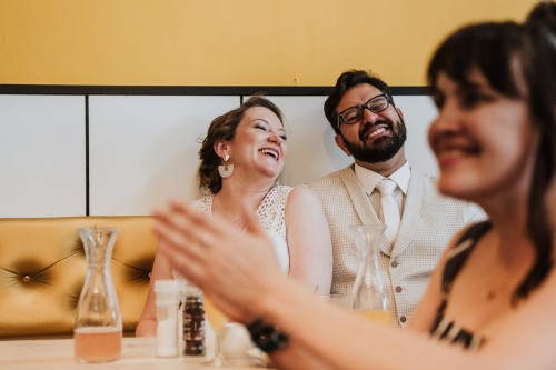 Ein Brautpaar sitzt an einer gelben Wand und lacht.
