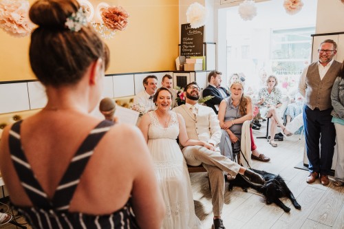 Eine Frau steht mit Mikrofon und ein Brautpaar mit Gästen sitzen im Raum und lachen.
