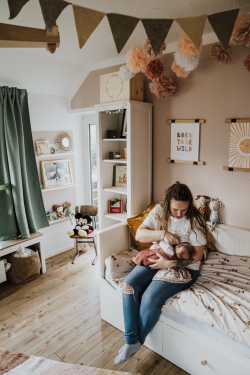 Eine Frau, die auf einem Bett im Kinderzimmer sitzt und ihr Baby hält.