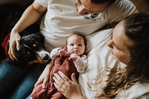 Die jungen Eltern halten ihr Baby in der Mitte und von der Seite schaut deren Hund