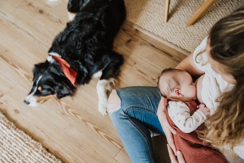 Eine Frau stillt ihr neugeborenes Baby und daneben liegt ein schwarzer Hund.