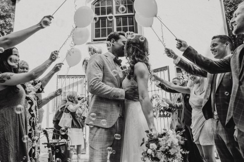 Ein schwarz-weißes Bild von einem küssenden Brautpaar und deren Gästen die einen Spalier mit Luftballons machen.
