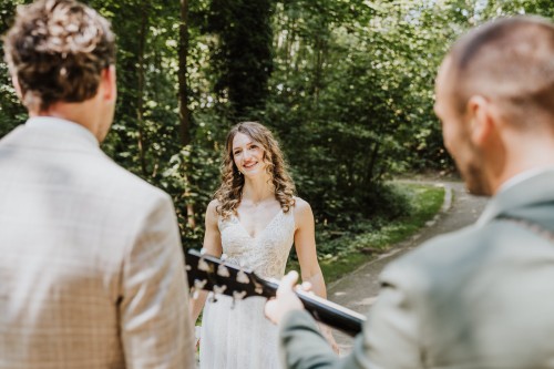 Ein Brautpaar steht im Wald und ein Mann spielt Gitarre.
