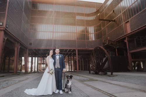 Ein Brautpaar steht mit einem Hund vor einer industriellen Kulisse.