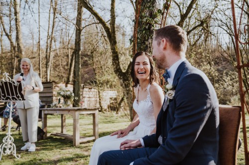 Eine Braut und ein Bräutigam sitzen vor einem Baum und lachen, im Hintergrund steht eine Frau und hält ein Mikro in der Hand.