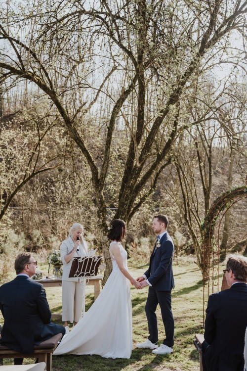 Ein Brautpaar steht vor einem Baum, eine Frau hinter denen hält eine Rede und die Gäste schauen zu.