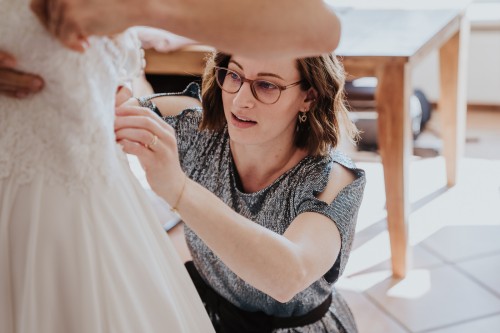 Eine Frau zieht der Braut das Brautkleid an.