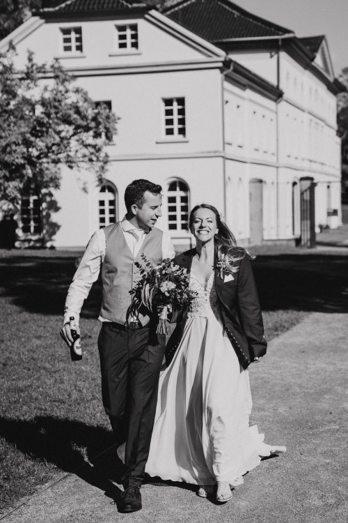 Eine Braut mit Sakko und ein Bräutigam mit Bier in der Hand gehen gemeinsam zum Standesamt.