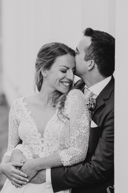 Ein schwarz-weißes Bild von einem Bräutigam der eine Braut am Kopf küsst.