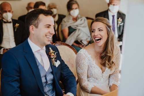 Eine Braut und ein Bräutigam sitzen im Standesamt und lachen herzlich.