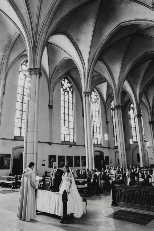 Ein Brautpaar steht in einer Kirche und die Gäste sitzen im Hintergrund.