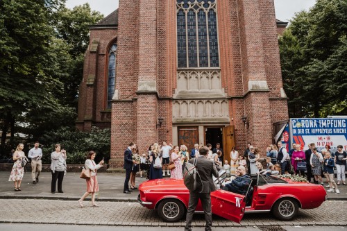 Eine Gruppe von Menschen und ein rotes Auto vor einer Kirche.