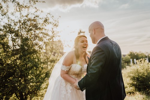 Eine Braut steht vor einem Bräutigam und lacht sehr herzlich.