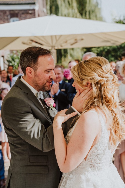 Ein Brautpaar umarmt sich und Gäste schauen im Hintergrund zu.